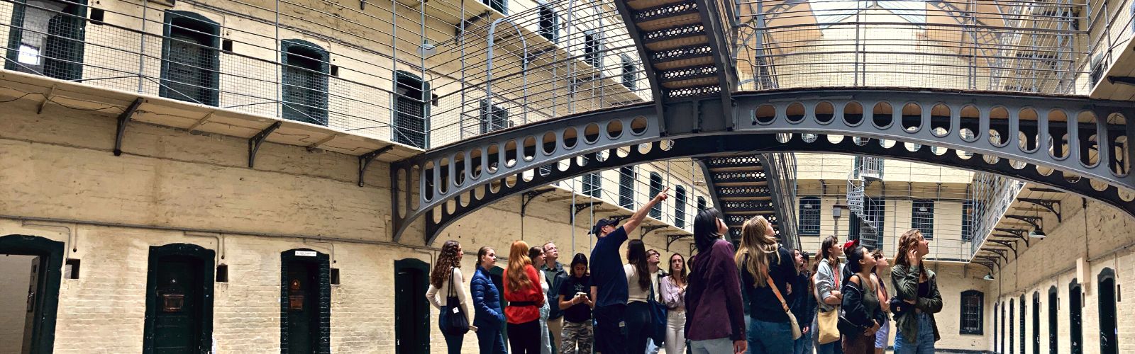 Students stare where a man is pointing inside a brick building with many metal archways.